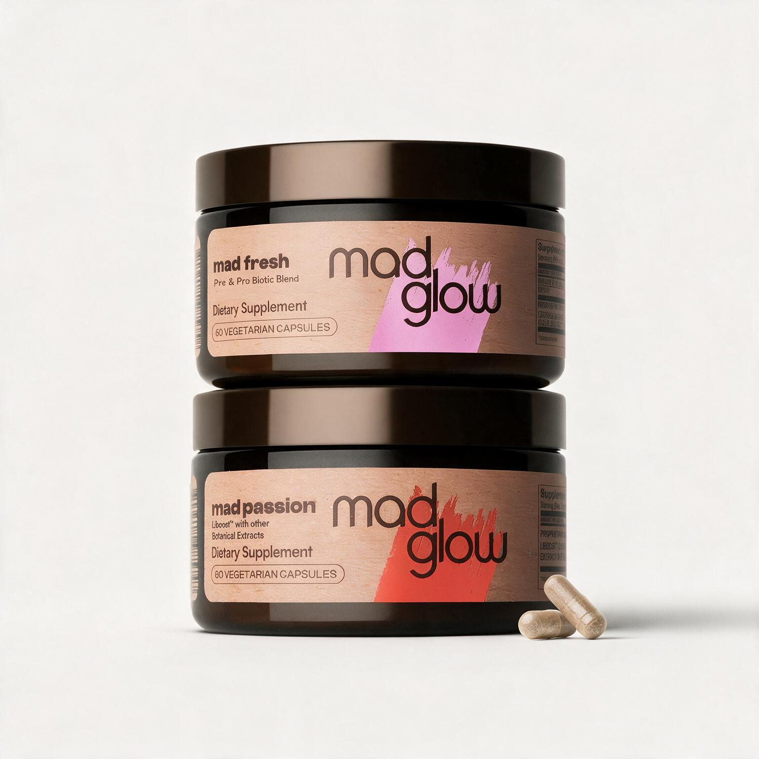 Two stacked dark amber supplement jars on a light gray background, with two capsules in front. The top jar label reads 'mad fresh' and the bottom jar label reads 'mad passion', both branded 'mad glow'.