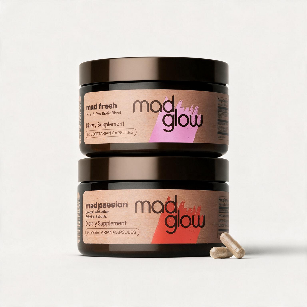 Two stacked dark amber supplement jars on a light gray background, with two capsules in front. The top jar label reads 'mad fresh' and the bottom jar label reads 'mad passion', both branded 'mad glow'.